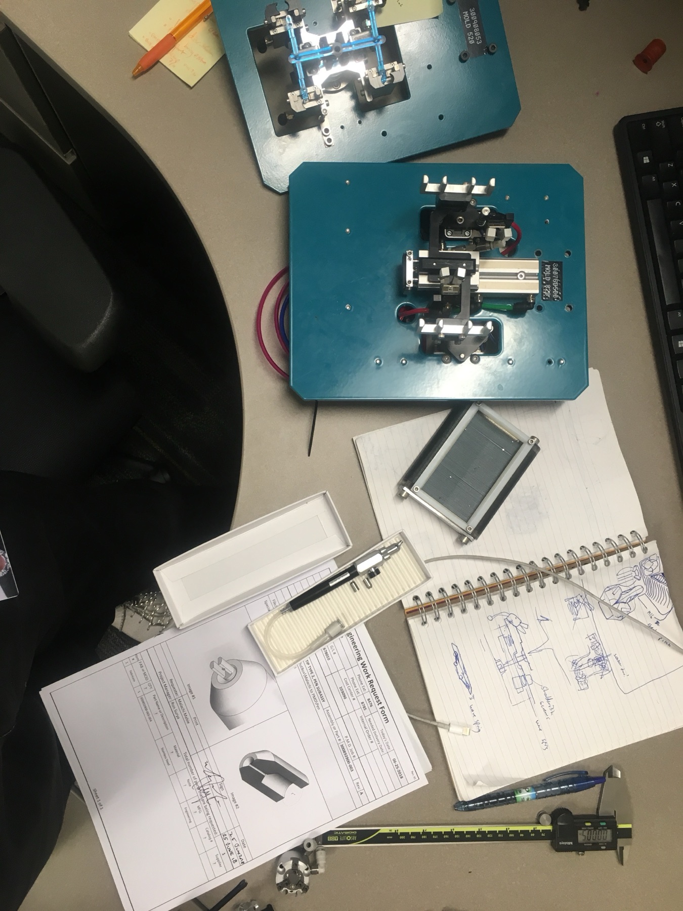 Pneumatic fixtures, drawings, calipers, and notes on a manufacturing workbench.